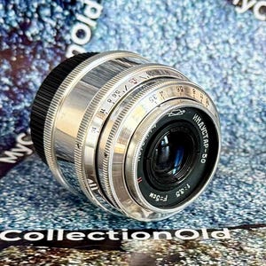 Puede incluir: Un objetivo de cámara plateado y negro vintage con el texto "ИНДУСТАР-50" y "1:3.5 F=5CM" visibles. El objetivo tiene anillos ajustables con números y marcas. Un accesorio de fotografía.