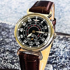 May include: A close-up of a vintage wristwatch with a black face and gold-colored casing. The watch features white numerals, red accents, and a brown leather band. The watch face displays the numbers 5, 10, 15, 20, 25, 30, 35, 40, 45, 50, and 55.