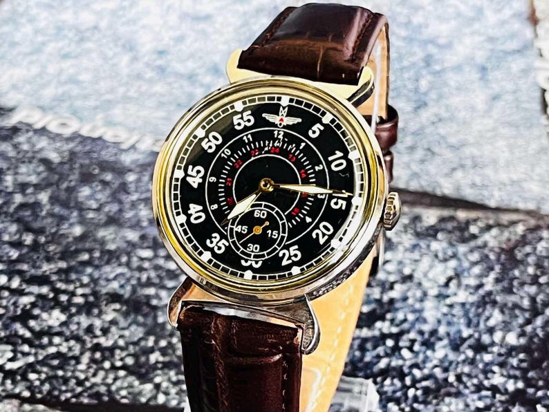 May include: A close-up of a vintage wristwatch with a black face and gold-colored casing. The watch features white numerals, red accents, and a brown leather band. The watch face displays the numbers 5, 10, 15, 20, 25, 30, 35, 40, 45, 50, and 55.