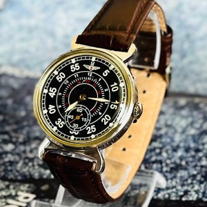 May include: A vintage-style wristwatch with a black face, white numerals, and a subdial. The watch has a gold-colored bezel and a brown leather strap. The watch face displays the numbers 5, 10, 15, 20, 25, 30, 35, 40, 45, 50, and 55.