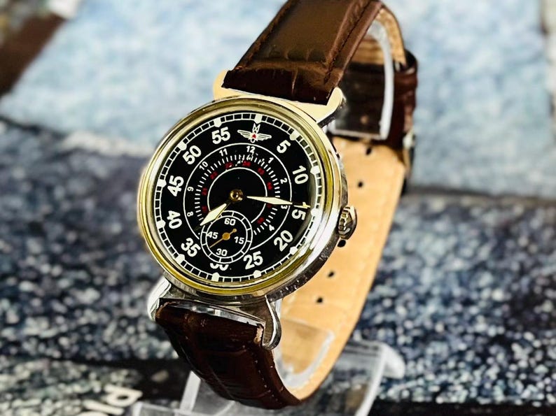 May include: A vintage-style wristwatch with a black face, white numerals, and a subdial. The watch has a gold-colored bezel and a brown leather strap. The watch face displays the numbers 5, 10, 15, 20, 25, 30, 35, 40, 45, 50, and 55.