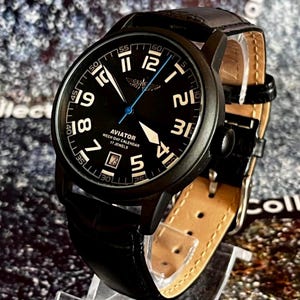 May include: Black Aviator watch with a black dial, white numerals, and a blue second hand. The watch features a leather band and the text "AVIATOR WEEK DAY CALENDAR 17 JEWELS".