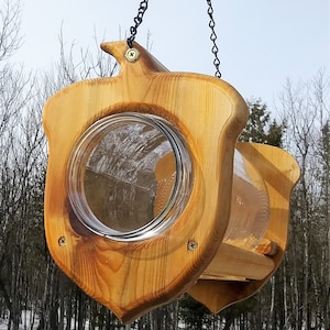 May include: A wooden bird feeder with a clear glass jar, shaped like a shield. The feeder is made of light-colored wood and is suspended by a black chain. The design includes a bird-like shape at the top.