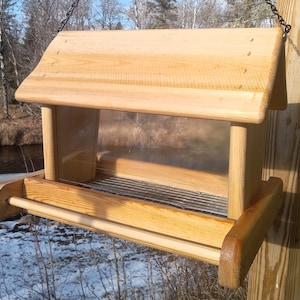 Op de afbeelding: Een houten vogelvoederhuisje met een transparante plastic voorkant en een metalen gaasbodem. De voederbak hangt aan een ketting en heeft een houten zitstok aan de voorkant.