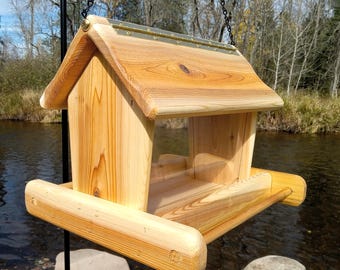 May include: A wooden bird feeder with a pitched roof and a tray. The feeder is made of light-colored wood and hangs from a chain. The design includes a clear plastic panel for viewing the birdseed.