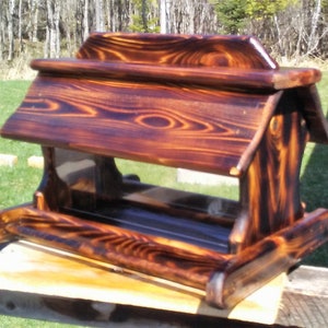 May include: A wooden bird feeder with a dark brown, almost black, wood stain. The feeder has a slanted roof and a clear plastic front panel. The feeder is sitting on a wooden surface.