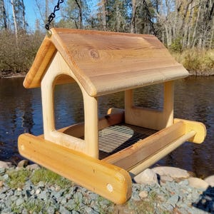 Puede incluir: Un comedero para pájaros de madera con techo inclinado, colgado de una cadena. El comedero tiene una base rectangular con un suelo de malla metálica y una pequeña percha. La madera es de color marrón claro con vetas visibles.