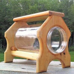 Könnte beinhalten: Ein Futterspender aus Holz für Vögel oder Eichhörnchen, aus Naturholz mit einem durchsichtigen Glasbehälter für Futter. Der Futterspender hat ein einfaches Design mit einer flachen Oberseite und einem erhöhten Sockel.