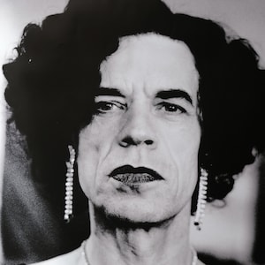 Op de afbeelding: Zwart-wit portretfoto van een persoon met donker krullend haar, die een lange parelketting draagt als oorbellen en een ketting. De persoon kijkt recht in de camera met een serieuze uitdrukking.