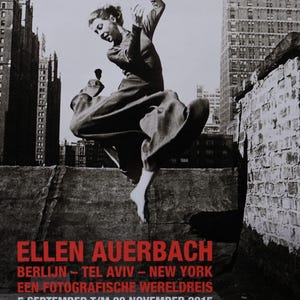 May include: Black and white photograph of a woman jumping in the air in front of a city skyline. The text "ELLEN AUERBACH" is in red letters at the bottom of the image. The text "BERLIJN - TEL AVIV - NEW YORK" is in black letters below the name. The text "EEN FOTOGRAFISCHE WERELDREIS" is in black letters below the cities. The text "5 SEPTEMBER T/M 29 NOVEMBER 2015" is in black letters below the world trip text. The text "MUSEUM DE FUNDATIE, ZWOLLE" is in black letters below the dates. The text "WWW.MUSEUMDEFUNDATIE.NL" is in black letters below the museum name.