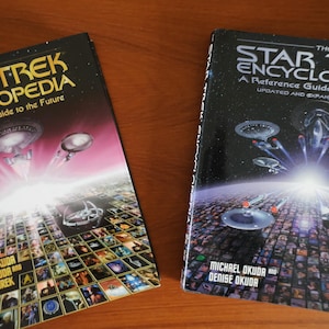 Puede incluir: Dos ejemplares de los libros "The Star Trek Encyclopedia". Uno tiene una portada rosa y blanca con naves espaciales y el otro una portada negra con naves espaciales. Ambos libros tienen el título y los nombres de los autores visibles.