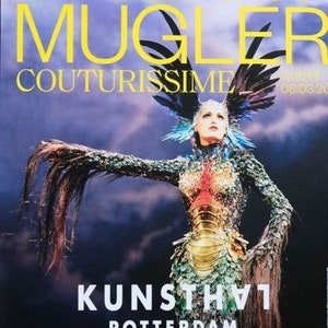 May include: A poster advertising a Thierry Mugler Couturissime exhibition at the Kunsthal Rotterdam. The poster features a model wearing a gold and green costume with a large feathered headdress. The text on the poster reads "Thierry Mugler Couturissime" and "Kunsthal Rotterdam".