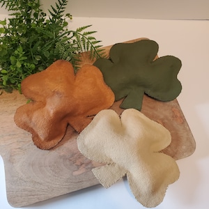 May include: Three felt shamrock shapes in different colors: brown, green, and beige. The shamrocks are on a wooden surface.