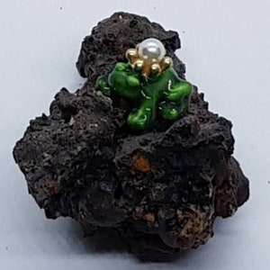 Puede incluir: Una pequeña figura de rana verde con una corona de flores de oro y perla sentada sobre una roca marrón oscura.