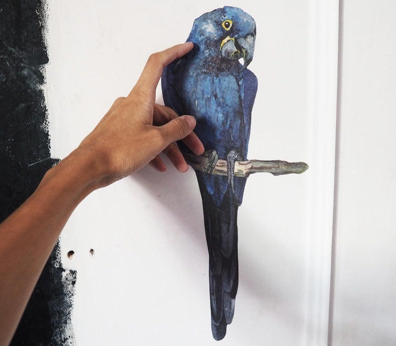 May include: A wall decal featuring a vibrant blue hyacinth macaw perched on a brown branch. The bird has deep blue feathers, a dark grey beak, and a bright yellow ring around its eye. The illustration is vertically oriented and applied to a wall.