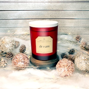 Könnte beinhalten: Eine rote Glas-Kerze mit weißem Deckel, beschriftet mit "dream" aus der "winter candle collection". Die Kerze steht auf einem Holzsockel, umgeben von dekorativen Ornamenten und Tannenzapfen. Die Kerze ist 283 Gramm.