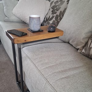 May include: A wooden side table with a dark metal frame. The table has a rectangular top with a natural wood finish. A smartphone, a mug, and a remote control are on the table. The table is next to a grey sofa with decorative cushions.