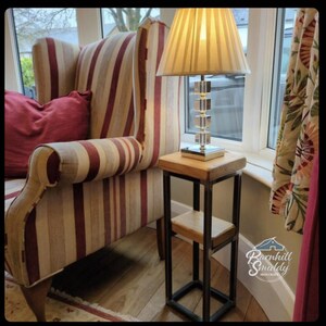 May include: A wooden side table with a metal frame and two shelves. The table is next to a striped armchair with a red cushion. A lamp with a cream-coloured shade sits on the table. The table is labelled "Roxhill Smiddy Iron Craft".