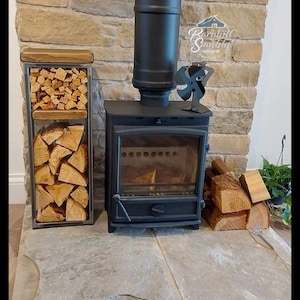 May include: A black metal wood-burning stove with a vertical chimney pipe, a small black stove fan on top, and a tall rectangular metal log rack filled with split firewood. The logo Barnhill Smiddy Iron Craft is visible on the stone wall.
