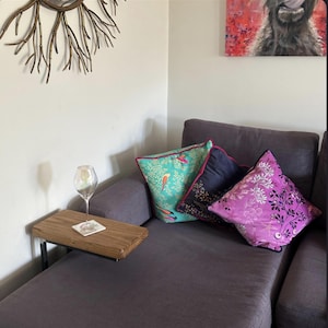 May include: A rectangular wooden armrest table with a dark metal frame, holding a clear glass wine goblet on a square coaster. The table is attached to a dark grey sofa featuring three decorative throw pillows in teal, navy, and magenta.