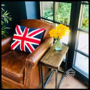 May include: A brown leather armchair with brass nailhead trim features a square pillow with a red, white, and blue Union Jack pattern. Beside it stands a rectangular wooden side table with a metal frame holding a glass vase of yellow flowers.