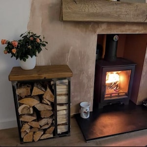 May include: A rectangular metal log storage rack with a dark wood top surface, filled with stacked split firewood and smaller kindling pieces. A white ceramic pot containing a small rose plant with orange flowers sits on the wooden top.