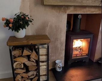 May include: A rectangular metal log storage rack with a dark wood top surface, filled with stacked split firewood and smaller kindling pieces. A white ceramic pot containing a small rose plant with orange flowers sits on the wooden top.