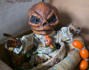 Könnte beinhalten: Eine handgefertigte Halloween-Puppe mit einem Kürbiskopf und einem strengen Gesichtsausdruck. Die Puppe trägt ein gemustertes Hemd und sitzt in einem geflochtenen Korb. Der Text "Hey, Pumpkid" ist sichtbar. Eine Kette aus orangefarbenen Perlen sorgt für einen dekorativen Touch.
