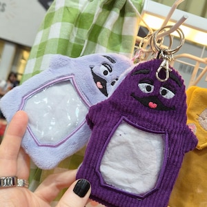 Puede incluir: Dos llaveros de peluche con forma de personajes de dibujos animados. Uno es lavanda, el otro es morado. Ambos tienen una ventana transparente y una cara sonriente bordada. Los llaveros cuelgan de un estante metálico.