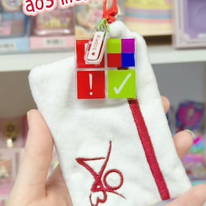 Puede incluir: Una bolsa blanca con un diseño bordado en rojo y un acento rojo. Adjunto hay un dije con cuadrados coloridos y una pequeña etiqueta que dice "Kudos". El texto "a03 merch? a03 merch!" está en el fondo.