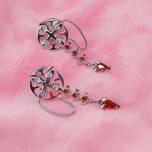 Puede incluir: Un par de pendientes de plata con un diseño colgante. Cada pendiente presenta un dije circular con un patrón en forma de cruz y cristales transparentes. Los dijes están conectados a una cadena con acentos de cristal rojo.