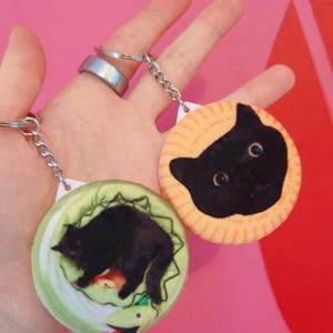 May include: Two round keychains with cat designs. One keychain features a black cat on a green background with a snake, the other has a black cat on an orange background. Both have silver keychains.