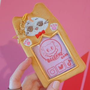 Puede incluir: Una funda de teléfono de felpa dorada con forma de gato con una ventana transparente para una foto. La funda tiene una pequeña cara de gato bordada con una pajarita y un charm de estrella. La funda también tiene iconos de redes sociales bordados: Instagram, Twitter y TikTok. El texto "BEEFANS.COM" está bordado en la funda.
