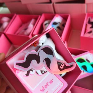 Peut inclure: Une boîte rose contenant un jouet en peluche et un autocollant avec un motif de ver. L'autocollant a un motif noir et blanc avec le chiffre 8. La boîte contient également une carte rose avec le mot "Worm" et "Series 01" imprimés.