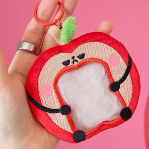 Peut inclure: Un porte-clés en forme de pomme rouge avec un centre blanc, des accents noirs et une feuille verte. Le porte-clés a un anneau rouge et un petit détail de feuille beige et verte. La pomme a un visage de dessin animé avec des joues roses.