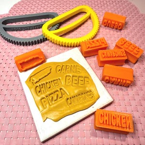 May include: A collection of orange food-themed stamps and cutters. The stamps include words like "CHICKEN", "BEEF", and "PIZZA". Two cutters are also present, one yellow and one gray, with a gear-like design. The items are on a pink surface.