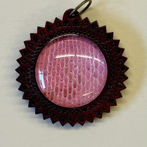 May include: A pendant with a burgundy-colored, starburst-shaped frame. The center features a pink, oval-shaped design with a scale-like pattern. A small, circular loop is attached to the top for hanging.