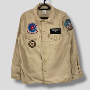 Peut inclure: Chemise boutonnée beige avec plusieurs écussons. Les écussons comprennent un emblème circulaire avec un oiseau et du texte, un écusson rectangulaire avec un motif d'aile et un écusson avec un motif rouge et bleu. La chemise a deux poches de poitrine et est suspendue à un cintre noir.