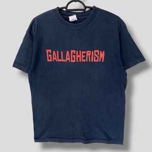 Puede incluir: Camiseta azul marino con la palabra "GALLAGHERISM" impresa en rojo en el pecho. La camiseta está colgada de una percha negra. Es una prenda de vestir informal.