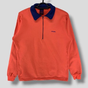 Puede incluir: Sudadera naranja de manga larga con cuello azul marino y cremallera de un cuarto. La sudadera tiene el nombre de la marca "Patagonia" bordado en azul marino en el pecho. La prenda está colgada de una percha negra.