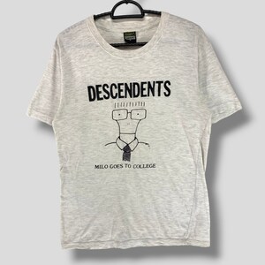 Puede incluir: Camiseta gris claro con la palabra "DESCENDENTS" en letras negras encima de un dibujo animado de un personaje con gafas y corbata. El texto "MILO GOES TO COLLEGE" está debajo.