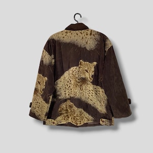 May include: A brown jacket with a leopard print design. The jacket has a dark brown collar and cuffs, and the leopard print is in shades of tan and brown. The jacket is hanging on a black hanger.