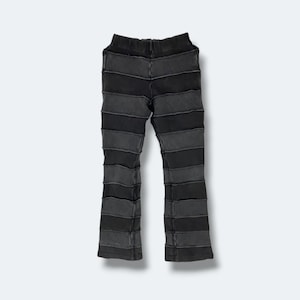 May include: Black and grey patchwork trousers with a flared leg. The trousers are constructed from horizontal strips of fabric in varying shades of grey and black, sewn together. The waistband is solid black.