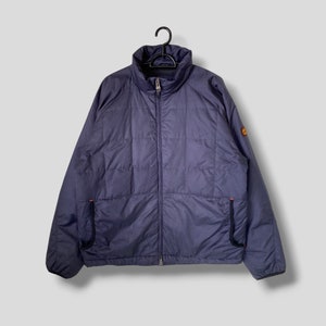 Puede incluir: Chaqueta acolchada azul marino con cuello alto y cremallera completa. La chaqueta tiene dos bolsillos delanteros y un pequeño logotipo en la manga superior derecha. Colgada de una percha negra.