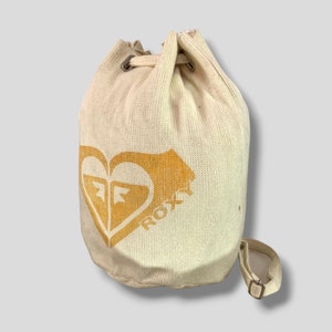 May include: Cream-colored drawstring bag with a gold heart-shaped logo and the word "ROXY". The bag has a shoulder strap and metal eyelets. The bag is made of a textured fabric.