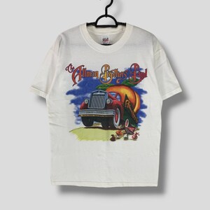 Może przedstawiać: Biała koszulka z grafiką czerwonej ciężarówki przewożącej dużą brzoskwinię, z napisem "The Allman Brothers Band" i latami "1969-1999". Koszulka wisi na czarnym wieszaku.