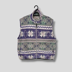 Puede incluir: Un chaleco de forro polar vintage con cremallera completa y cuello alto. El chaleco presenta un estampado geométrico en tonos morado, verde, gris y blanco. Cuelga de una percha negra sobre un fondo gris claro.