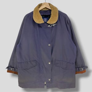 May include: A blue jacket with a corduroy collar and brown accents. The jacket has a button-up front, two front pockets, and adjustable cuffs. The jacket is hanging on a wooden hanger.
