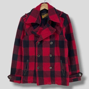 Op de afbeelding: Een rood-zwarte geruite wollen jas met een dubbele rij knopen. De jas heeft een reverskraag, twee voorzakken en manchetten met knopen. De jas hangt aan een houten kleerhanger.