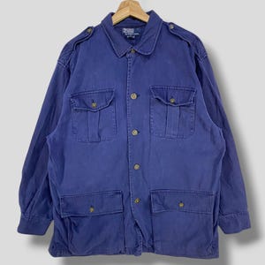 May include: A vintage blue jacket with four front pockets and button closures. The jacket has a collared neckline and shoulder epaulettes. The Polo Ralph Lauren label is visible on the back of the collar. The jacket is hanging on a wooden hanger.